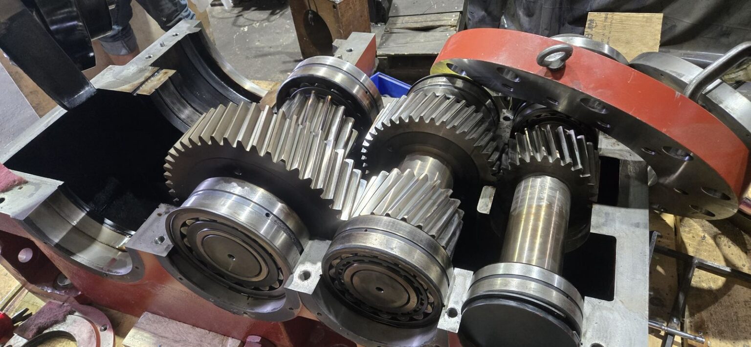 Twin-screw extruder gearbox opened for inspection, showing the helical gear train and bearings in the housing