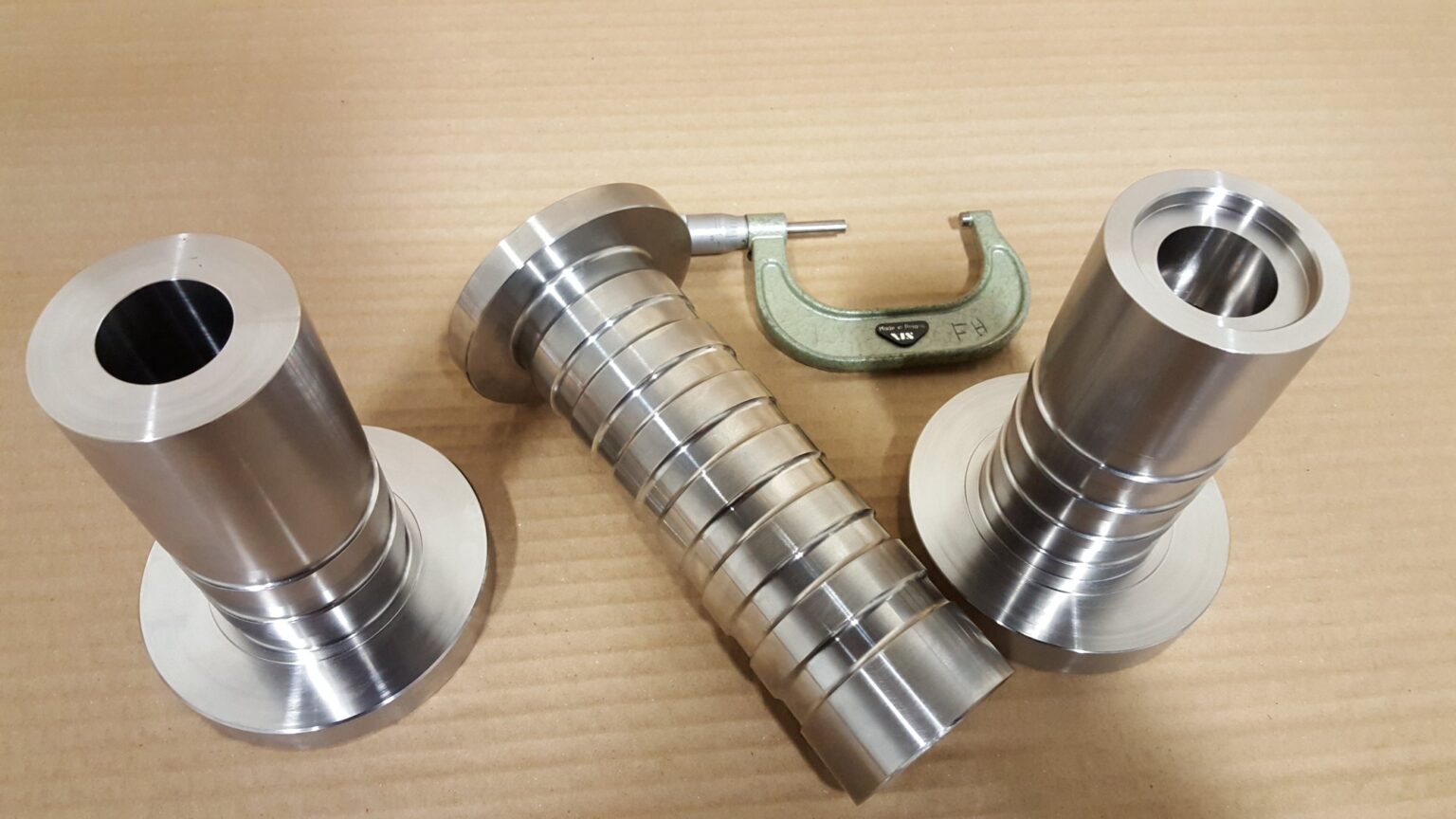 Three machined stepped sleeves with a micrometer alongside, grooved bodies and flanged bases