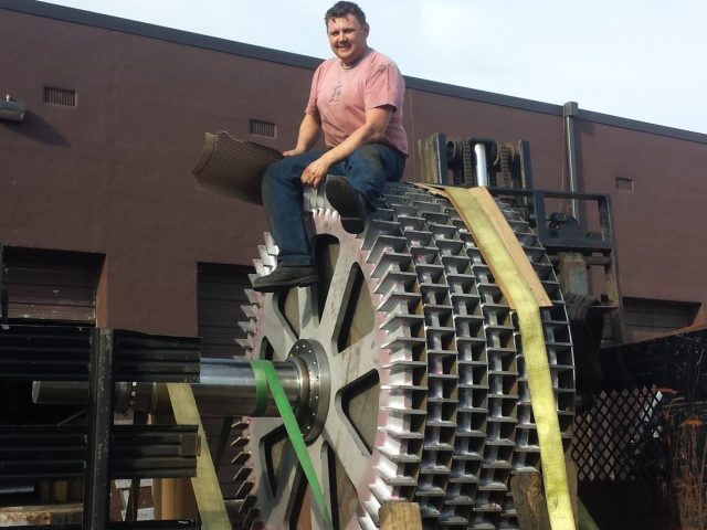Large spoked spur gear strapped to the flatbed, a man seated on the rim for scale